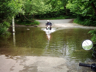 A motorcycle splashing through a ford surrounded by trees